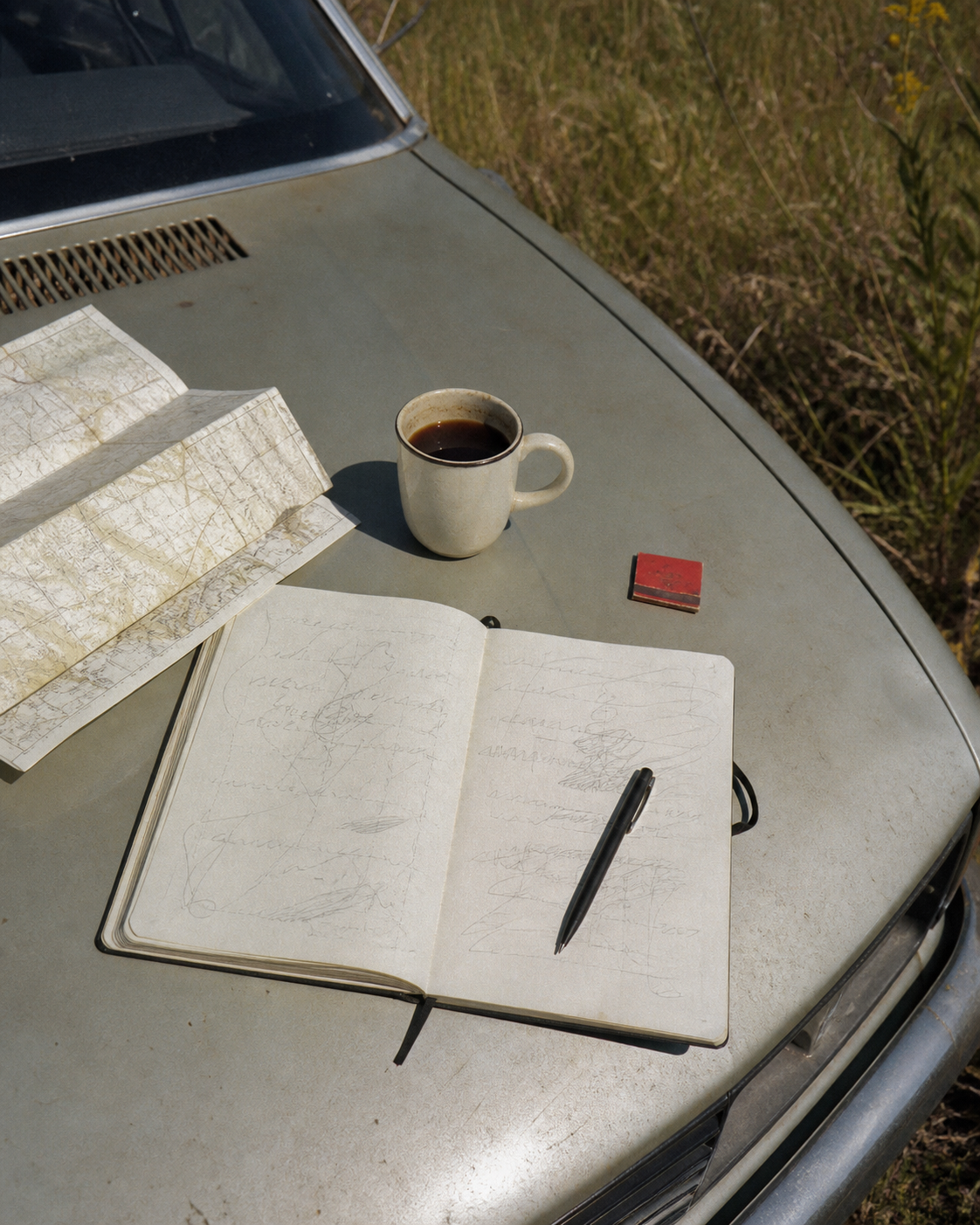 An open notebook, map, coffee, and pen on an old car hood